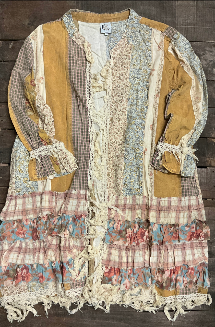 Mustard and sage patchwork duster with crochet lace front, plaid panels, lace-tied ruffle cuffs, and a tiered fringed ruffle hem, laid flat on dark wood