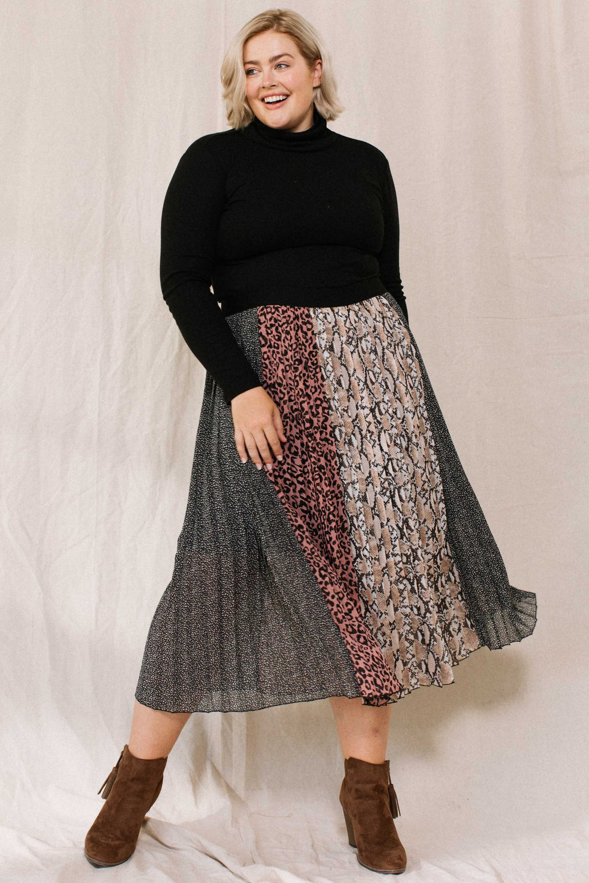 Model wearing a pleated midi skirt with leopard, snake and dot print panels, a black turtleneck and brown suede booties