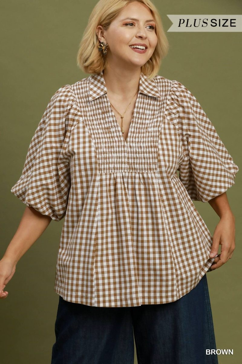 Plus size model wearing a brown gingham blouse with puff sleeves, split notch collar, and shirred yoke, paired with navy trousers