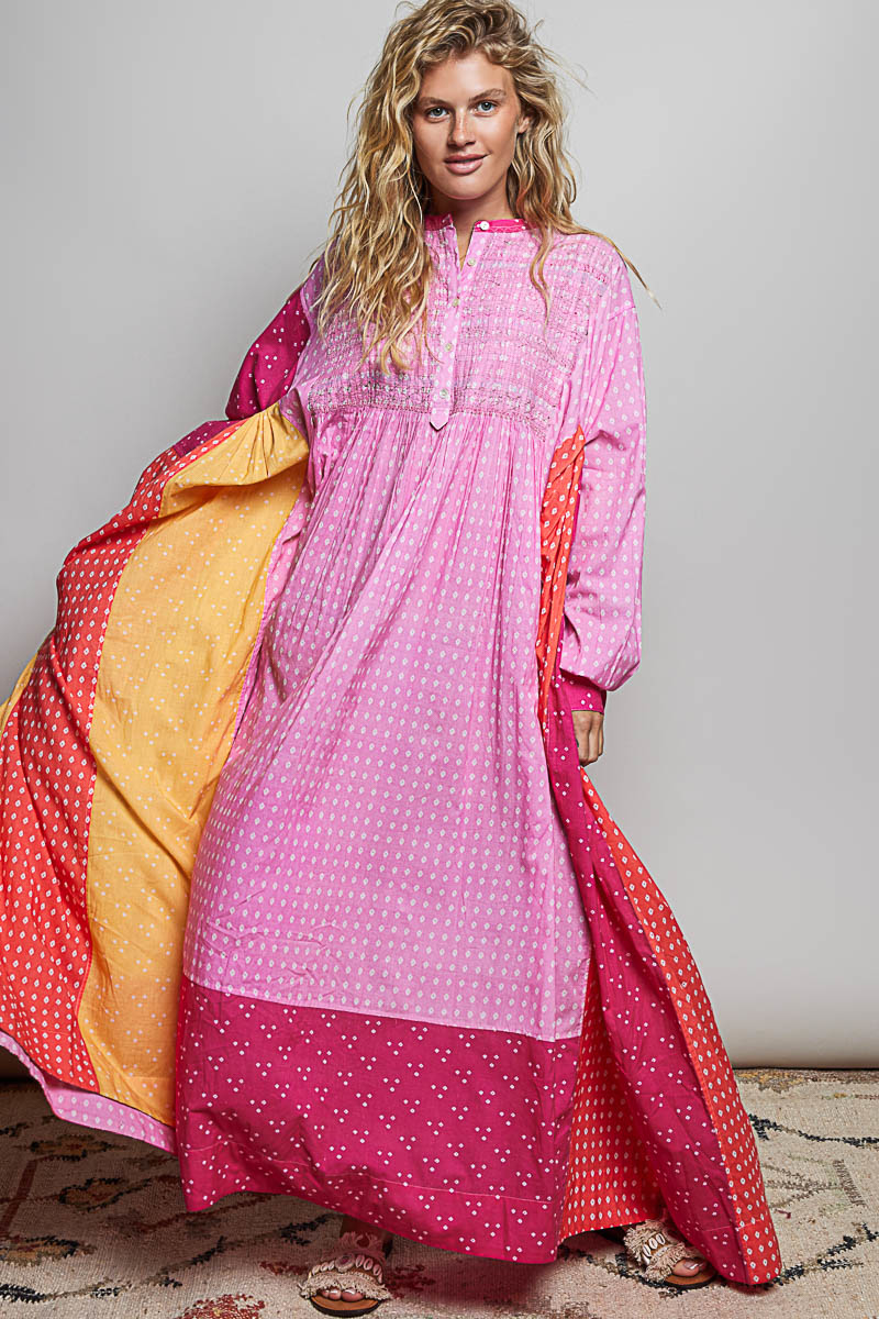 Model wearing a pink polka dot maxi dress with long sleeves, keyhole neckline, and layered flowing panels in pink, orange, and yellow