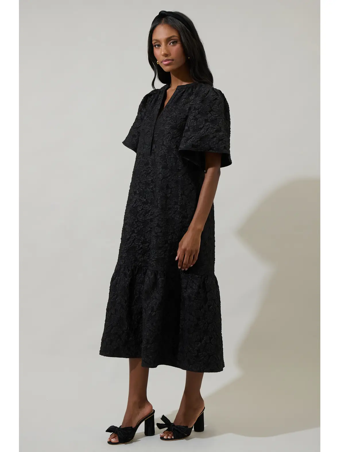 Model wearing a black textured midi dress with a split neckline, wide short sleeves, and a gathered lower tier.
