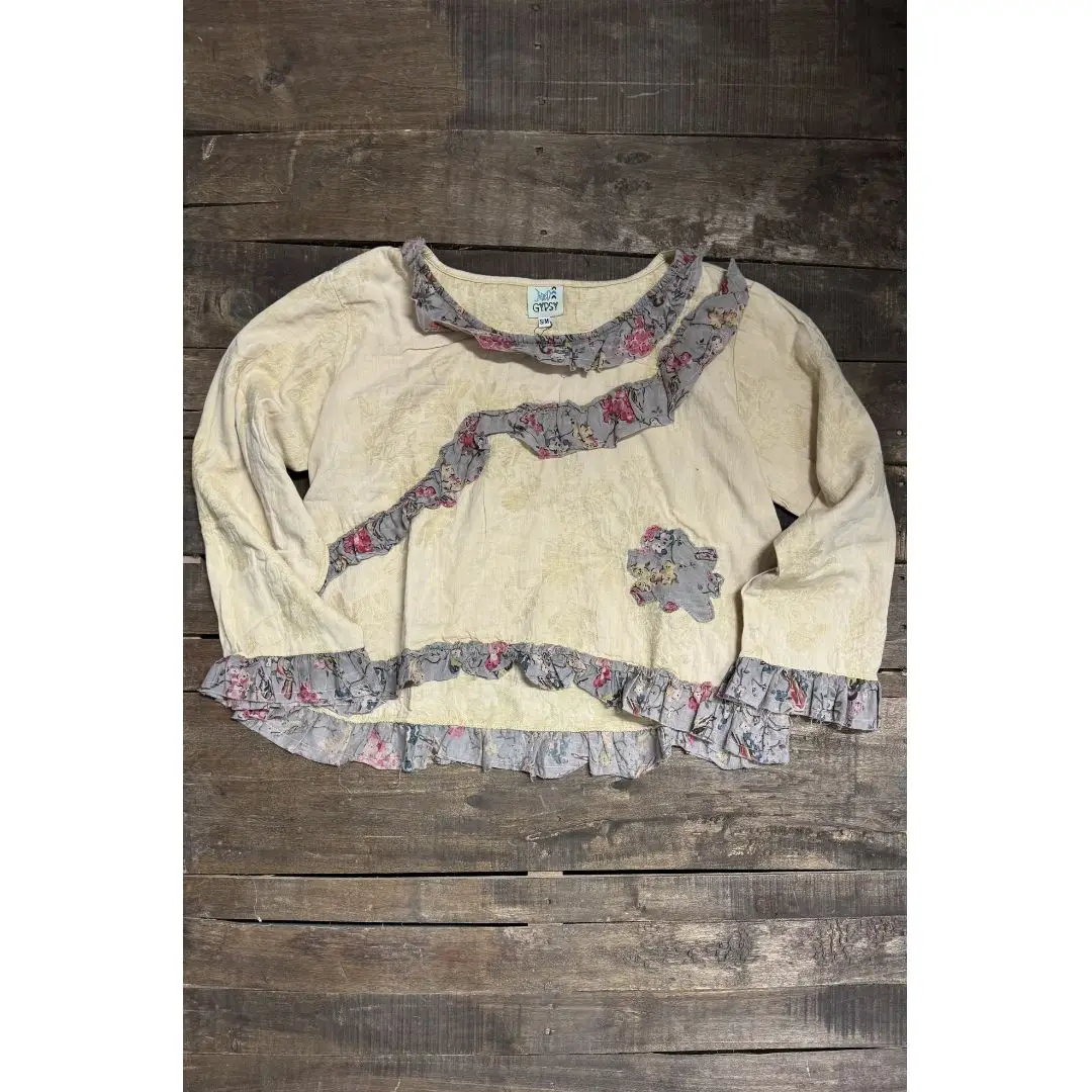 Cream long sleeve top with grey-lavender floral ruffle trim at the neckline, cuffs, and hem, and a flower-shaped floral appliqué on the chest