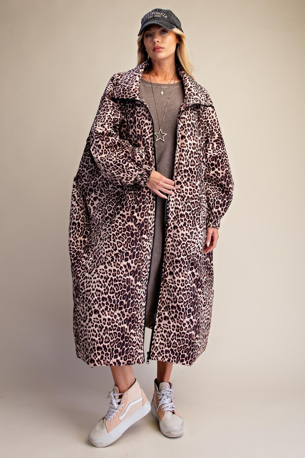Model wearing a cream and black leopard print longline puffer coat with a high collar, styled with a grey tee dress, sneakers, and a cap