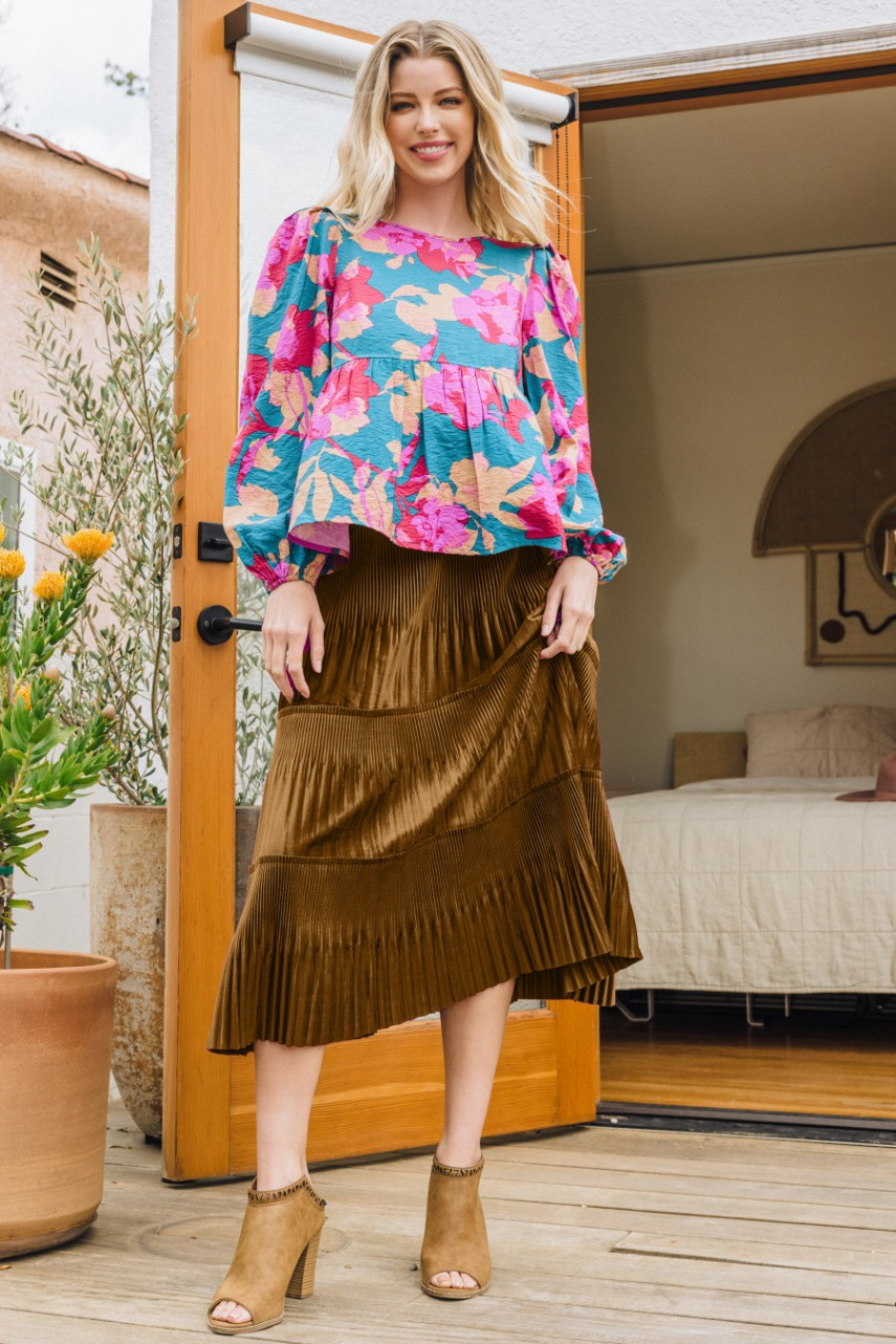 Model wearing a tiered pleated satin midi skirt in olive brown with a floral blouse and tan heeled booties