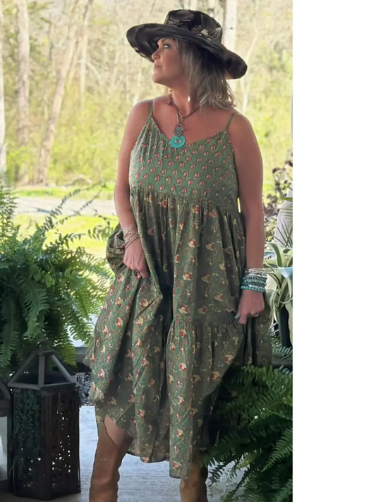 Model wearing an olive green ditsy floral tiered midi dress with spaghetti straps and side pockets, styled with a black wide-brim hat, turquoise jewelry, and brown boots