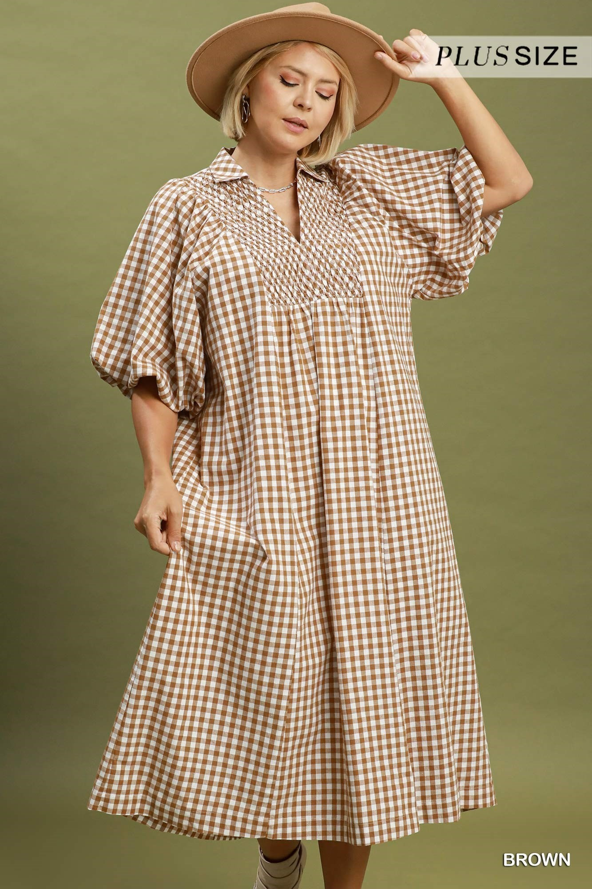 Plus size model wearing a brown gingham puff sleeve midi dress with smocked neckline detail and side pockets, styled with a tan hat