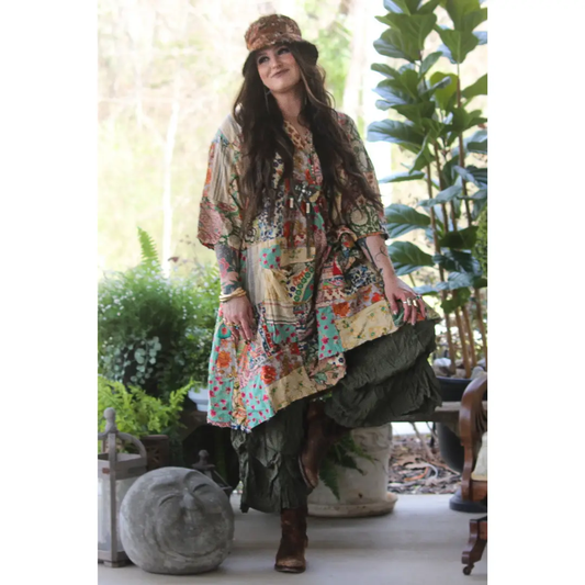 Woman wearing a flowy multicolor floral patchwork tunic dress with wide billowy sleeves and an asymmetric hem, styled with a wide-brim hat, layered necklaces, and boots